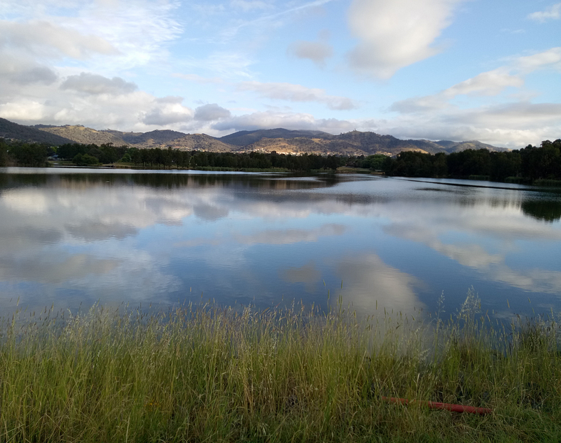 A serene lake reflecting the sky, surrounded by grassy fields and distant mountains.